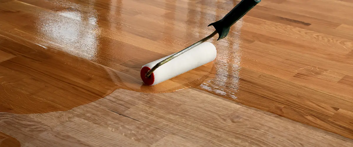 Applying a clear coat finish to hardwood floor with a roller, enhancing its natural color and adding a glossy protective layer.