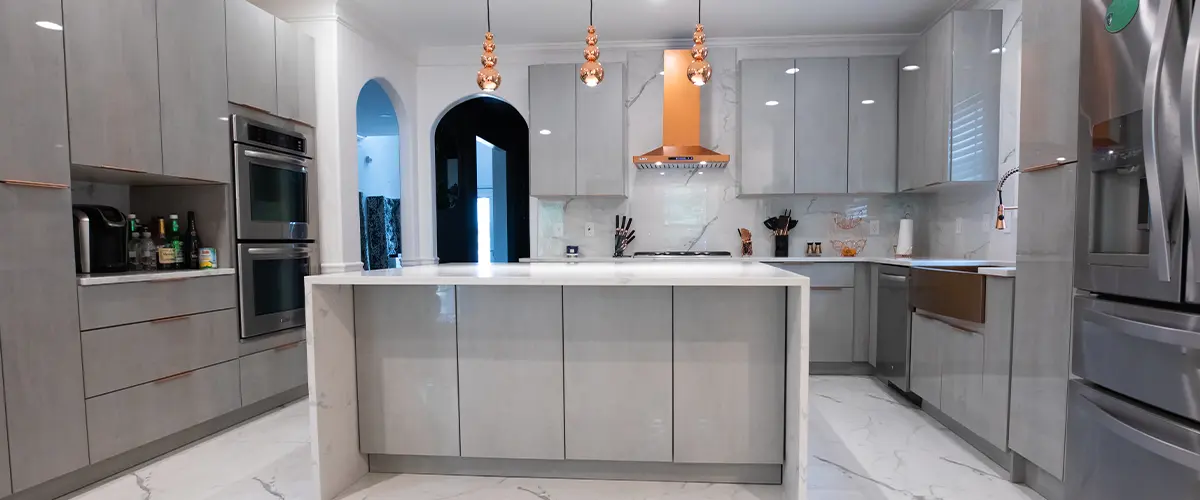 Modern kitchen with gray cabinetry, white marble countertops, and island, featuring pendant lights and stainless steel appliances.