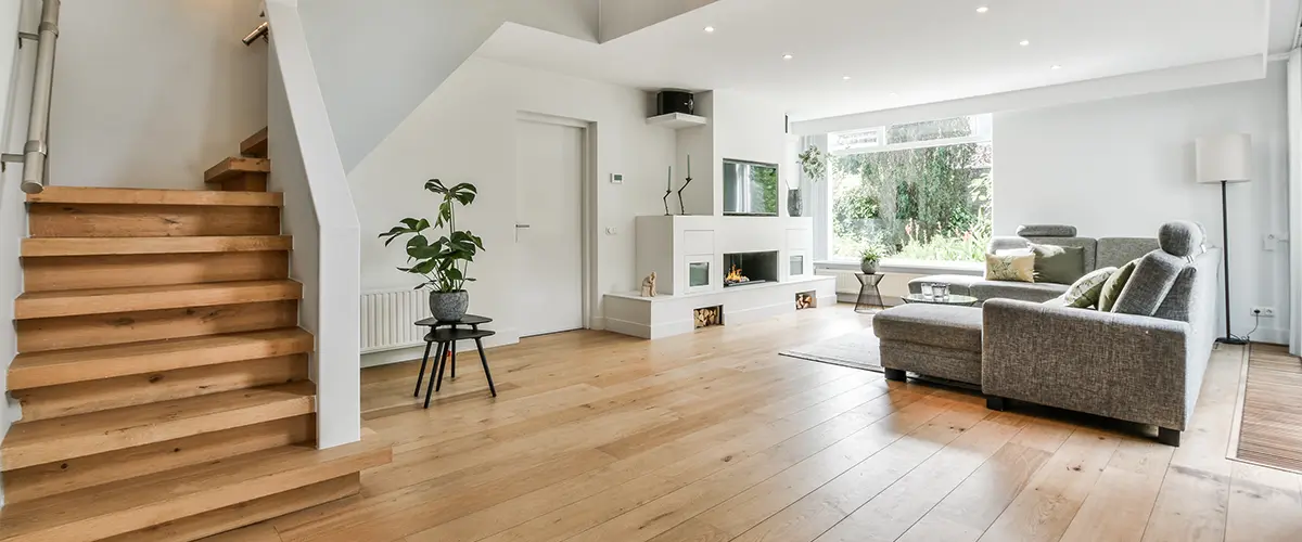 Spacious living room with light oak hardwood floors, modern staircase, gray sectional sofa, and large windows letting in natural light.