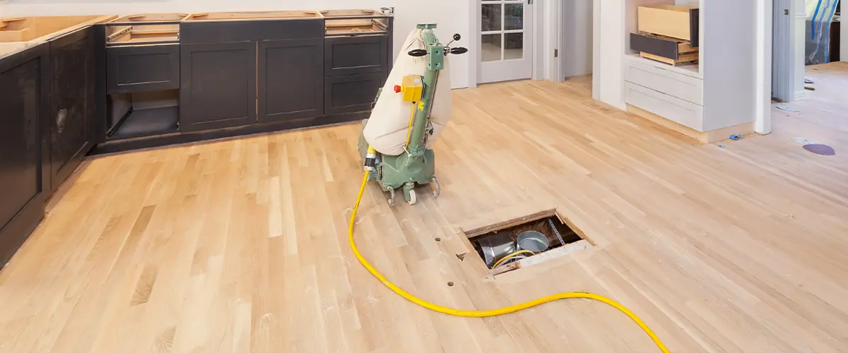 Under-construction kitchen with hardwood floor refinishing process,, black and gray cabinets, a floor sander, and a cutout for utilities.