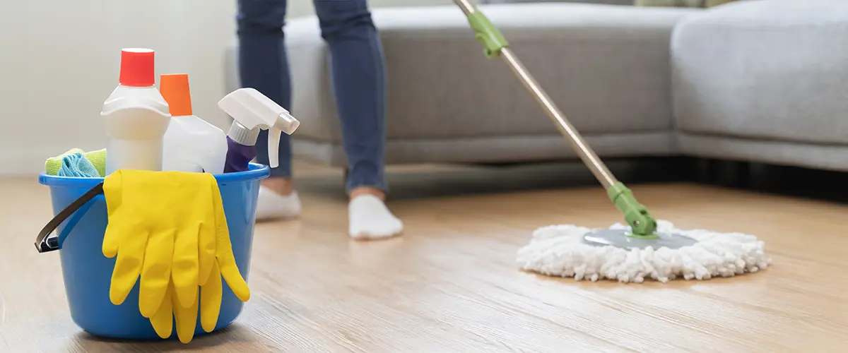 Close-up of a blue cleaning bucket with supplies and a person mopping a hardwood floor