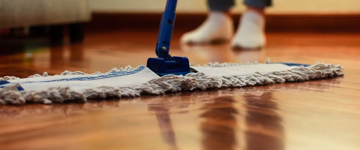 Person mopping a wooden floor with a blue mop, reflecting light from the floor