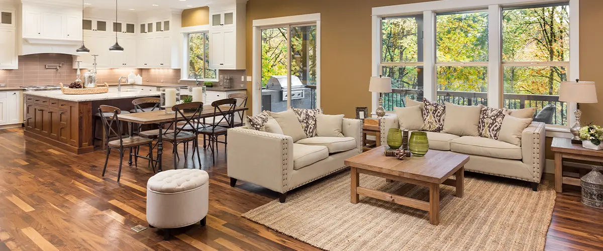 Modern open-concept living room and kitchen with hardwood flooring, beige sofas, wooden coffee table, and large windows.