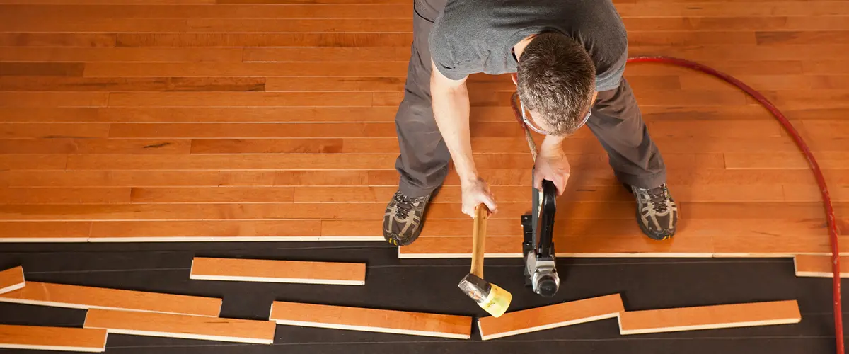 Professional installer laying hardwood flooring using a mallet and nail gun, showcasing a home improvement project in progress.